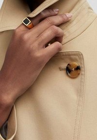 Tissu de trench-coat beige avec un col large, orné d’un grand bouton en écaille. Main affichant une bague dorée et noire à un doigt.