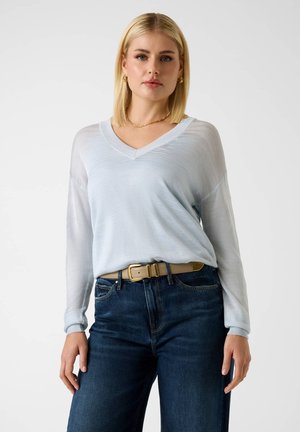 Blondynka o prostych włosach, ubrana w jasnoniebieski sweter z dekoltem w serek, ciemnoniebieskie dżinsy, beżowy pasek oraz złotą biżuterię, stojąca na białym tle.