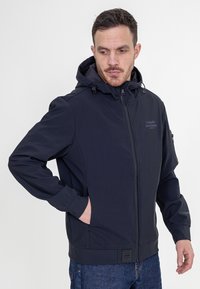 Marineblauwe softshell jack met capuchon, voorrits en zijzakken. Beschikt over een textuurstof en een logo op de borst met verstelbare koordjes.