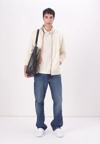 Giacca a zip crema con accenti arancioni, polo beige, jeans blu e sneaker bianche. Una borsa a spalla nera completa l'outfit.