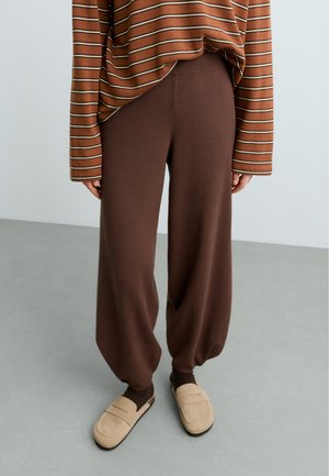 Personne portant un pantalon ample marron, des chaussures beige à enfiler et un haut marron à rayures manches longues, debout sur un sol gris contre un mur clair.
