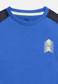 Μπλε αθλητική μπλούζα με στρογγυλή λαιμόκοψη, με μαύρες πλαϊνές επιφάνειες στους ώμους και ανοιχτό κίτρινο λογότυπο Puma στο στήθος και μέσα στο γιακά.