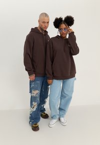 Brown hoodies with front pockets, paired with distressed blue jeans. One has round glasses; the other features a tattoo and multicolored shoes.