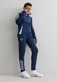 Zēns zila Arsenal treniņtērpā un baltās kedās stāv, smaidīdams pie teksturētas baltas sienas, ar roku tuvu sejai.