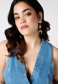 Jonge vrouw met lang, donker haar, gekleed in een denim top en grote, decoratieve oorbellen. Zachte uitdrukking met neutrale make-up tegen een witte achtergrond.