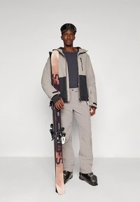 Χειμερινή στολή σκι που περιλαμβάνει γκρι ανοιχτή ζακέτα με μαύρες λεπτομέρειες, γκρι παντελόνι και μαύρα παπούτσια σκι· κρατάει ροζ και μαύρα πέδιλα με δεσίματα.