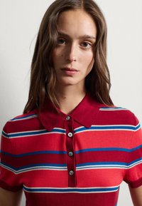 Rotes Poloshirt mit blauen und weißen Streifen, kurzen Ärmeln und einer Knopfleiste. Hergestellt aus weichem Stoff mit glatter Textur.