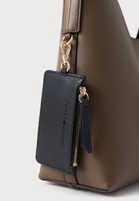 Ruda dirbtinės odos rankinė su tekstūruotu paviršiumi, turinti nuimamą juodos odos etiketę, auksinius priedus ir užtrauktuko kišenės detalę.