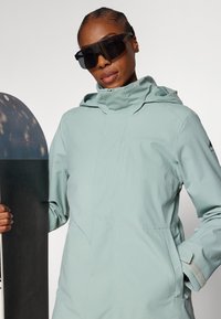 Lys teal ski-jakke med hette, glidelås og justerbare mansjetter; har en glatt tekstur og bæres med sorte solbriller og et snowboard.