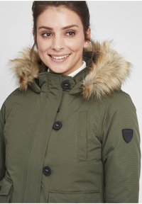Olive green parka jacket with a fur-lined hood, black buttons, and a logo patch on the sleeve. Features a zip and snap closure.