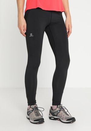 Leggings de sport noirs en matériau extensible, avec un design ajusté et un petit logo. Associés à des chaussures grises et noires.