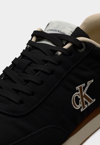 Μαύρα αθλητικά παπούτσια με υφασμάτινο επάνω μέρος, μπεζ κορδόνια, λαστιχένια σόλα και κεντημένο λογότυπο "CK" σε καφέ χρώμα στο πλάι. Μινιμαλιστικός σχεδιασμός.