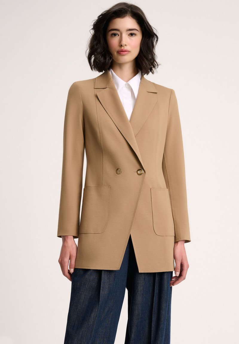 Beige double-breasted blazer with two front pockets, structured lapels, and a straight cut, paired with dark blue wide-leg trousers.