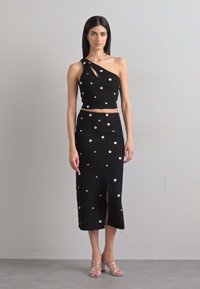 Tenue noire en deux pièces comprenant un crop top asymétrique avec découpe et une jupe midi avec fente avant, toutes deux ornées de petites appliques florales.