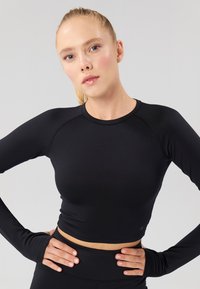 Zwarte crop top met lange mouwen, voorzien van een aansluitend ontwerp, een gladde textuur en duimgaten in de mouwen. Subtiel logo nabij de zoom.