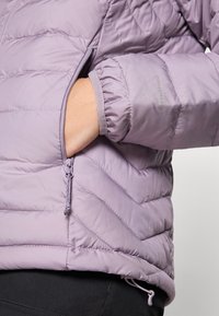 Lys lilla isolert jakke med chevron-søm, med en glidelås lomme og elastiske mansjetter, laget av et glatt og mykt materiale.