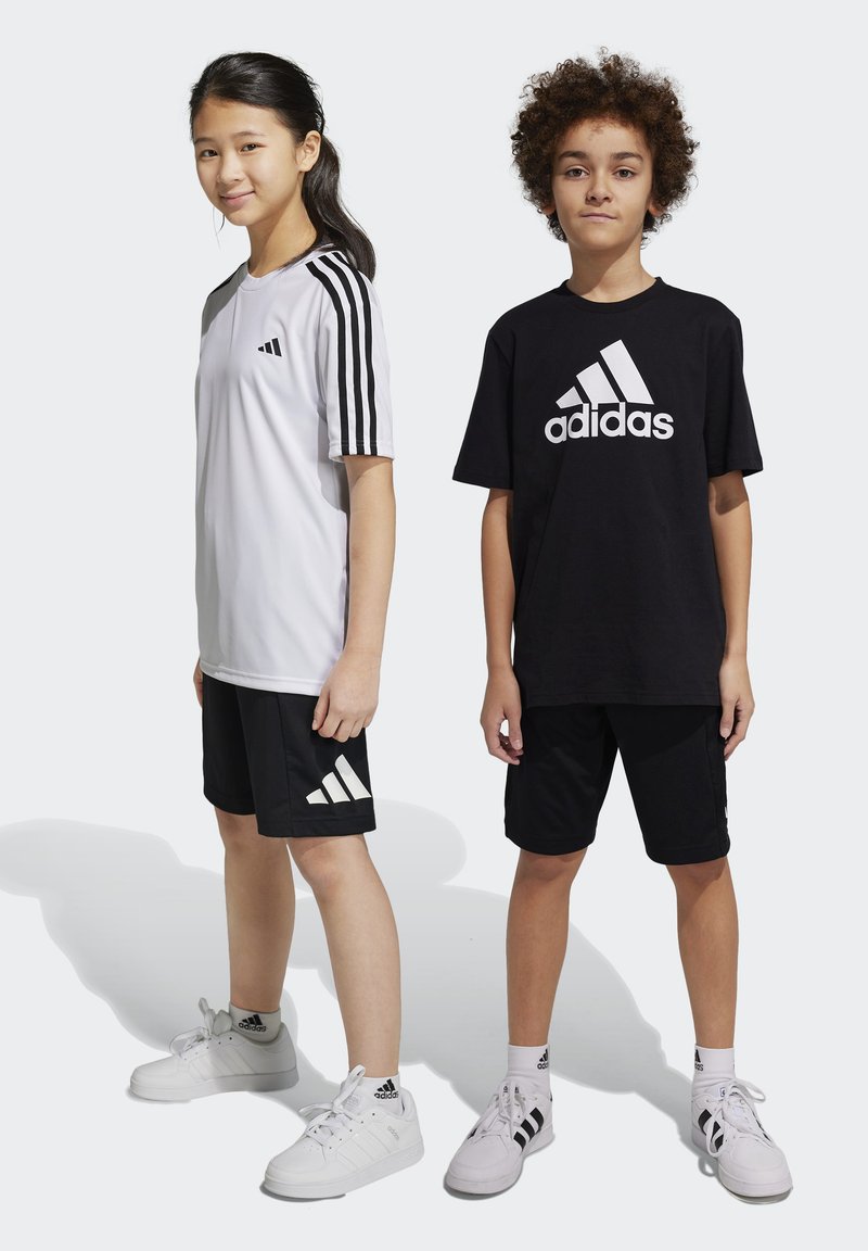 Dos niños llevan ropa deportiva: una camiseta blanca de Adidas con rayas negras y pantalones cortos negros, y una camiseta negra de Adidas con un logo blanco y pantalones cortos.