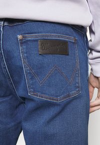 Džinsai su tamsiai mėlynu atspalviu, rudos odos etikete su užrašu "Wrangler" ir išskirtiniu zigzaginiu siuvimu ant užpakalinių kišenių.