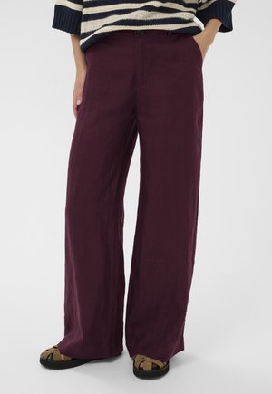 Žena stojaca s jednou rukou v vrecku, oblečená vo fialových širokých nohaviciach, pruhovanom svetri a hnedých sandáloch s odhalenými prstami.