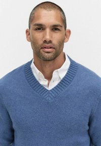 Niebieski sweter w serek z dzianiny, z prążkowanym kołnierzykiem, noszony na białej koszuli z kołnierzykiem, o fakturowanej tkaninie i luźnym kroju.