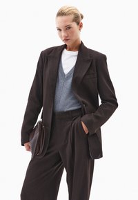 Blazer ajustado marrón sobre un chaleco de suéter gris, camiseta blanca y pantalones a juego. Se sostiene un bolso de cuero marrón con una mano.
