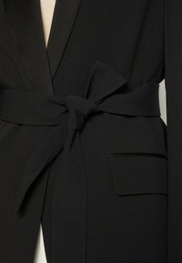 Blazer noir sur mesure avec une large ceinture en tissu nouée en nœud à la taille et une poche à rabat sur le côté droit.
