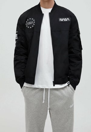 Hombre con chaqueta negra de la NASA sobre camisa blanca y pantalones deportivos grises con las manos en los bolsillos, de pie frente a un fondo liso.