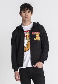 Crna zip-up dukserica s prednjim džepovima, s crtežom crnog medvjedića na prsima i bijelom majicom s grafikom ispod.