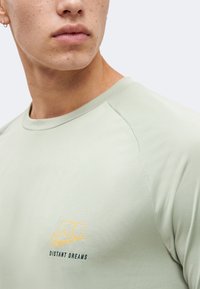 Κοντινό πλάνο ενός ατόμου που φοράει ανοιχτοπράσινο πουκάμισο με κίτρινο και μαύρο λογότυπο "NJ DISTANT DREAMS" στο στήθος, εμφανίζοντας το κάτω μέρος του προσώπου και το λαιμό.