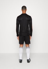 Man som står med ryggen mot kameran, iklädd en svart långärmad sportströja, svarta shorts med orange ränder, vita knähöga strumpor och svarta fotbollsskor.