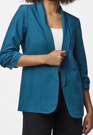 Frau trägt einen türkisfarbenen Blazer über einem weißen Top, mit einer Hand, die den Blazer richtet, gezeigt von den Schultern bis zu den Hüften.