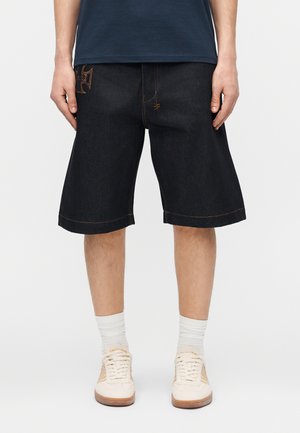 Άνδρας που φοράει φαρδιά μαύρα τζιν σορτς, λευκές κάλτσες και μπεζ αθλητικά, στέκεται με χαλαρά τα χέρια δίπλα στο σώμα μπροστά σε ουδέτερο φόντο.