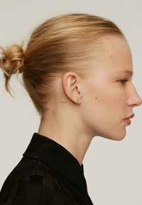 Vista de perfil de una persona con un moño rubio y elegante, vistiendo una camisa negra con cuello. Un pequeño tatuaje rojo con la palabra "oui" es visible detrás de la oreja.