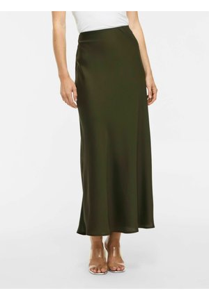 Femme portant une jupe taille haute, vert foncé, longueur cheville avec un haut blanc et des sandales transparentes à talons hauts, debout.