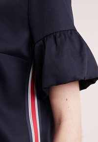 Blusa azul marino con mangas con volantes, con acentos de rayas verticales rojas y blancas a lo largo del costado. Textura de tela suave.
