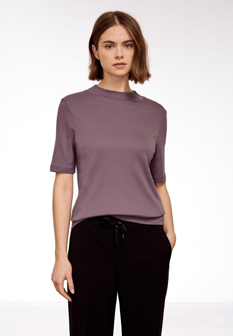 Jeune femme aux cheveux bruns courts portant un haut mauve à manches courtes et un pantalon noir, debout avec une main dans la poche devant un fond blanc uni.