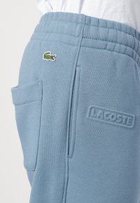 Lyseblå joggebukser laget av myk stoff. Har et brodert grønt krokodillemerke og et teksturert "LACOSTE"-merke på siden.