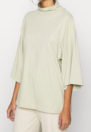 Kvinde iført en luftig lys grøn bluse med lange ærmer og høj krave samt off-white ribbukser mod en ensfarvet baggrund.