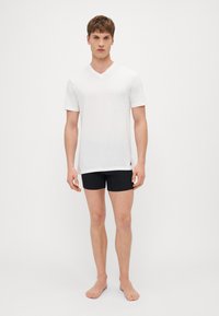 Weißes kurzärmliges V-Ausschnitt-T-Shirt aus weichem Stoff, kombiniert mit schwarzen eng anliegenden Shorts, Modell steht barfuß vor einem einfarbigen Hintergrund.