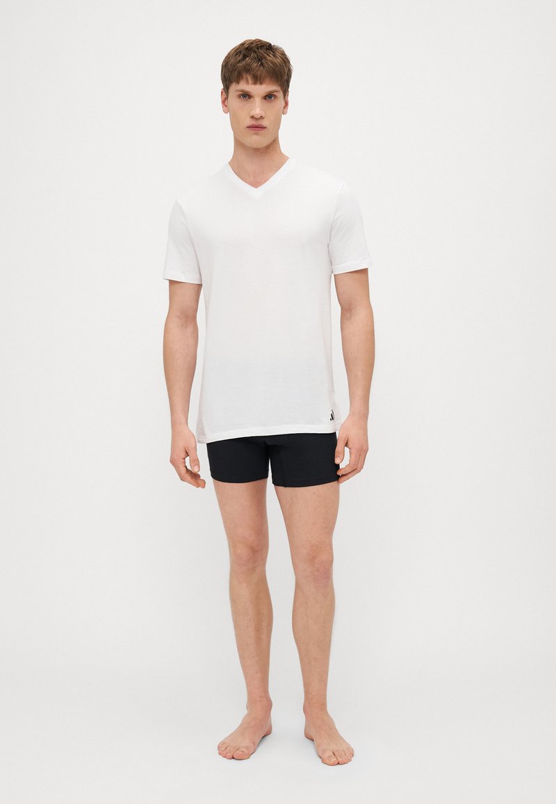 Weißes kurzärmliges V-Ausschnitt-T-Shirt aus weichem Stoff, kombiniert mit schwarzen eng anliegenden Shorts, Modell steht barfuß vor einem einfarbigen Hintergrund.