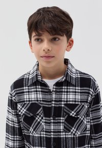 Camisa de franela a cuadros en blanco y negro con cierre de botones en la parte delantera, dos bolsillos en el pecho y un cuello doblado. Diseño de textura suave.