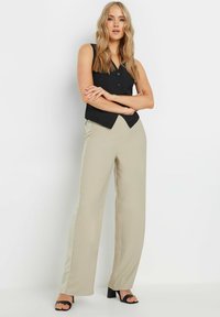 Un gilet sans manches noir avec des boutons, associé à un pantalon large beige clair. Le modèle porte des sandales à talons noires, mettant en valeur un design simple.