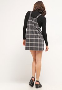 Robe salopette à carreaux gris avec bretelles ajustables et silhouette évasée, associée à un top noir ajusté à manches longues et à des sandales à talons épais.