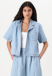 Camicia a maniche corte a righe blu in tessuto leggero, dotata di colletto, taschino frontale e chiusura con bottoni. Si abbina a pantaloni blu simili.
