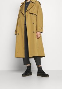 Beige trenchcoat i slätt tyg med dubbla knappar och axelregister, bärs över en grå topp och mörka byxor, i kombination med svarta stövlar.