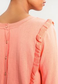 Růžový pletený svetr s volánkovými akcenty na ramenou a zapínáním na zádech na kulaté kovové knoflíky. Jemná textura a přiléhavý střih.