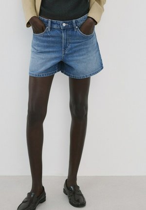 Kobieta ubrana w jasnoniebieskie szorty dżinsowe, beżową kurtkę, czarny top i czarne mokasyny, z rękami w kieszeniach, stoi na tle jednolitego białego tła.