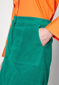 Personne portant une chemise orange à manches longues avec la main dans la poche d'une jupe en velours côtelé vert sarcelle, munie d'une fermeture éclair à l'avant et d'une grande poche avant.