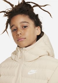 Chaqueta acolchada beige con cuello alto, cierre de cremallera y logo de Nike en blanco, con una textura suave y un diseño acolchado.