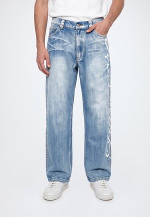 Le modèle porte un jean bleu clair à jambes larges, orné de coutures latérales décoratives, une chemise blanche et des baskets blanches. Une main repose dans une poche.
